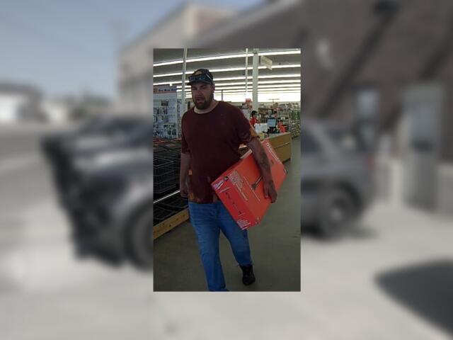 Bearded man in a burgundy shirt and cap carries a red-orange box through a store aisle.
