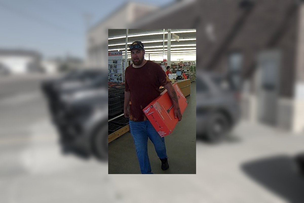 Bearded man in a burgundy shirt and cap carries a red-orange box through a store aisle.